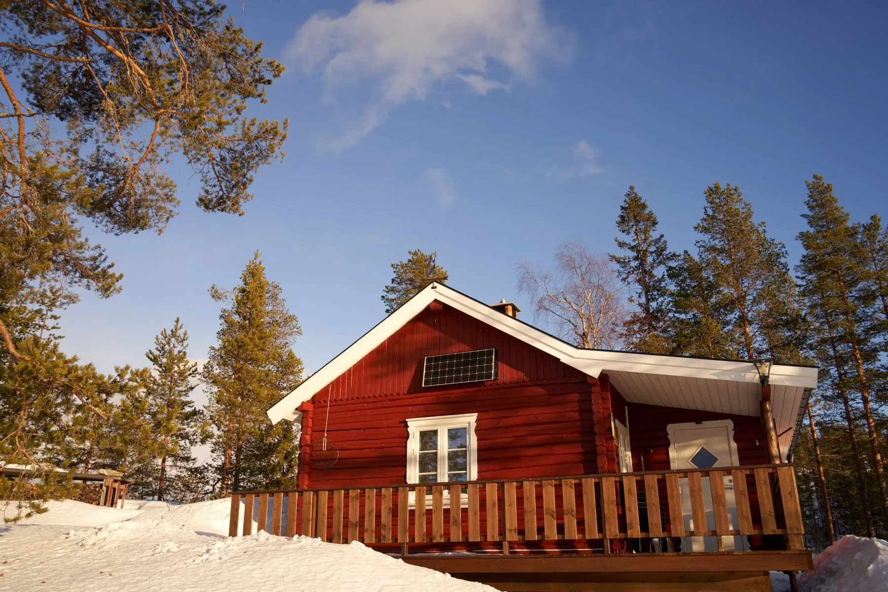 A red wooden cabin among snow-laden pines with a single solar panel catching low winter sun