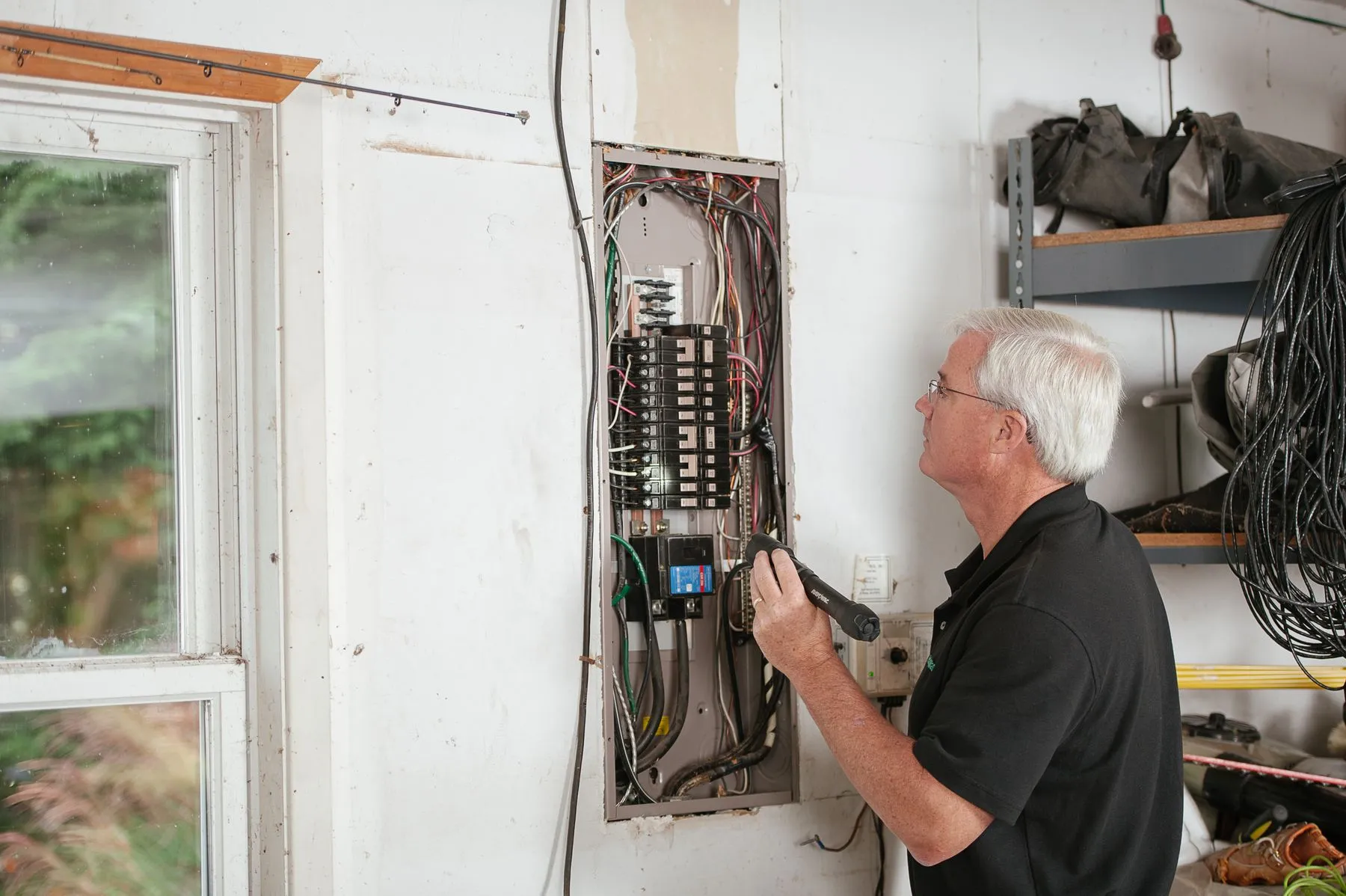 An electrician holding a flashlight while inspecting an open residential breaker panel on a garage wall