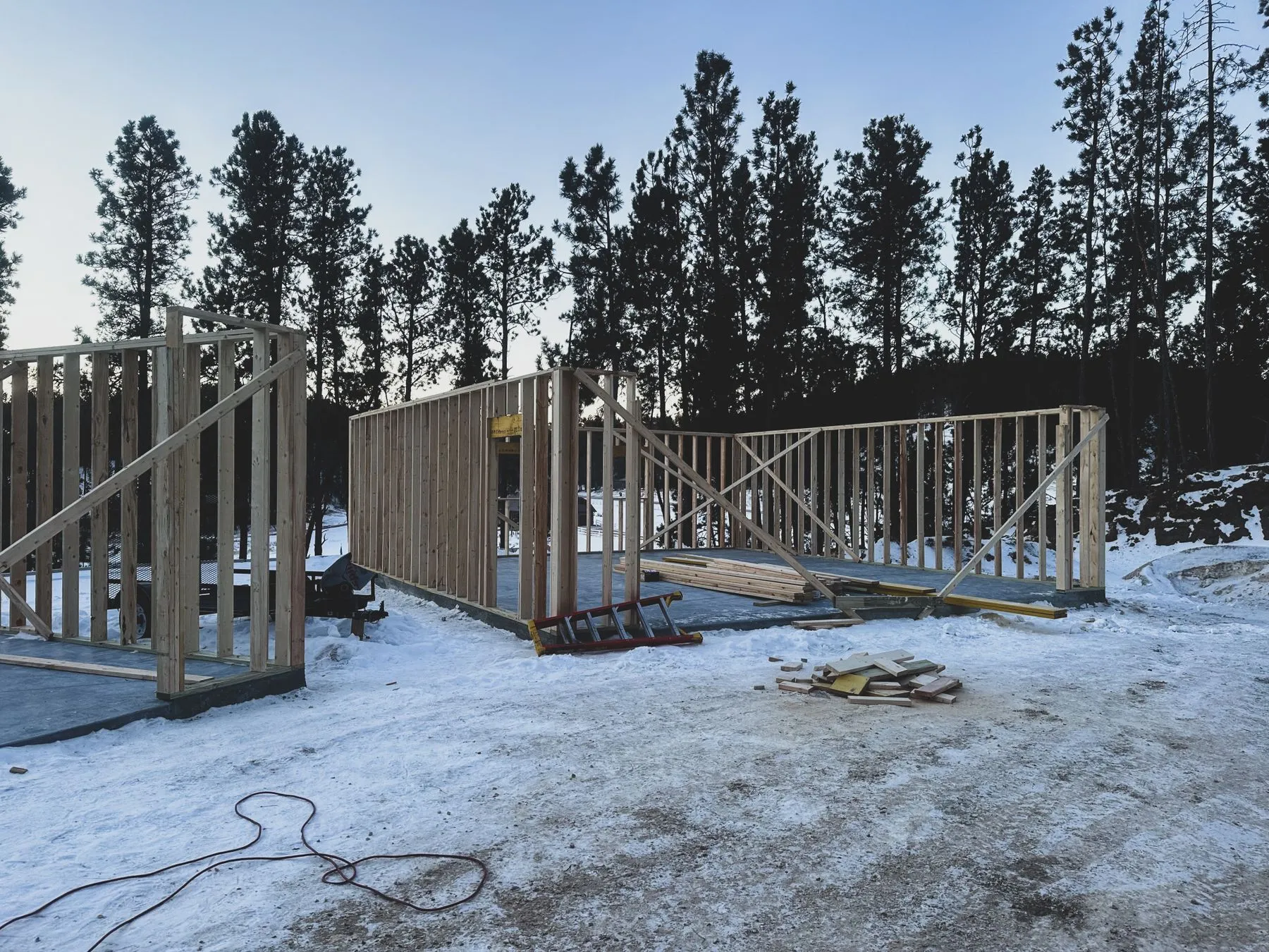 Wood-framed walls standing on a concrete slab at a snowy building site, with lumber stacked inside and pine trees behind