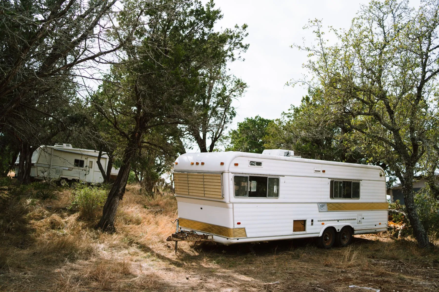 Two older white travel trailers parked among oak trees and dry grass on a rural Texas property