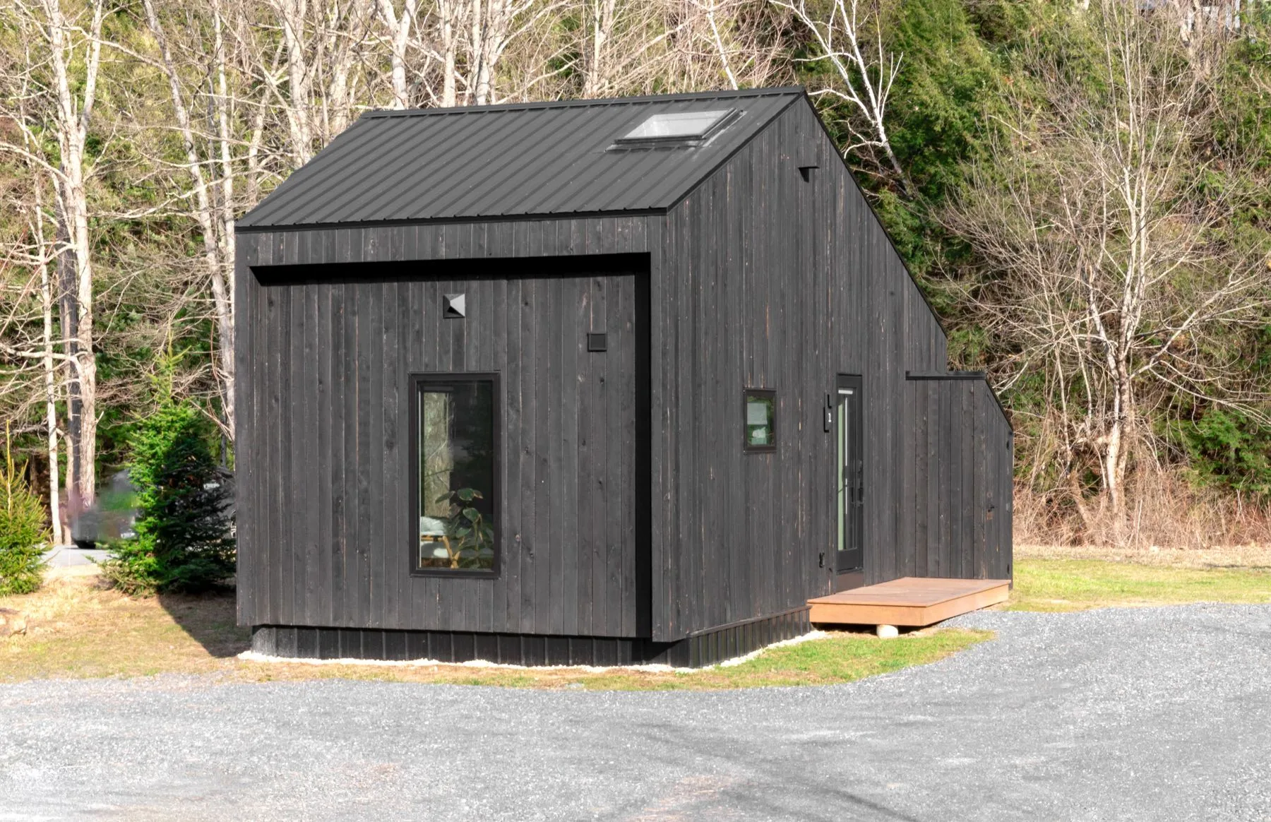 A small dark-sided tiny house with a metal roof and a skylight, set on a gravel pad at the edge of a wood